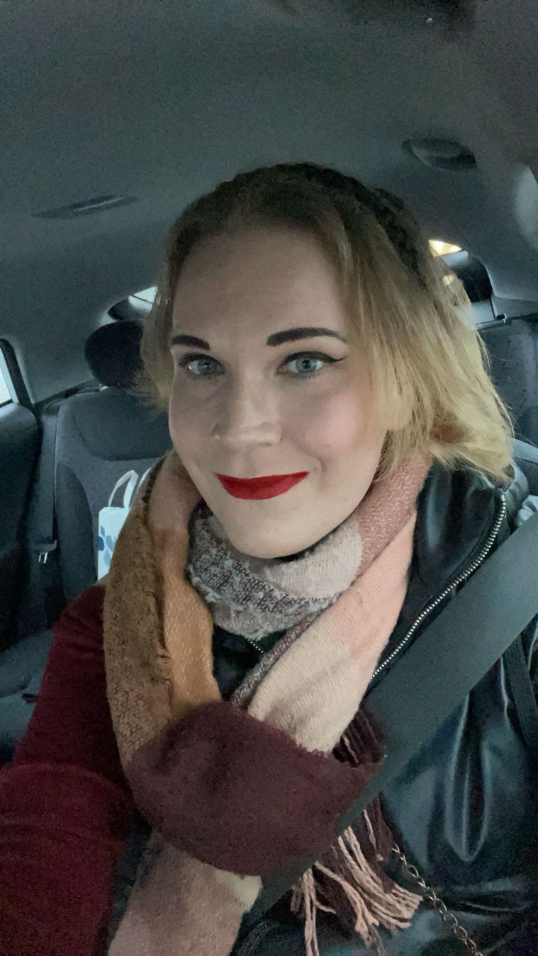 It's a selfie of me. With light brown hair and bold red lipstick smiles while seated in a car, wearing a black jacket and multiple scarves. The background includes the car's interior and back seat.