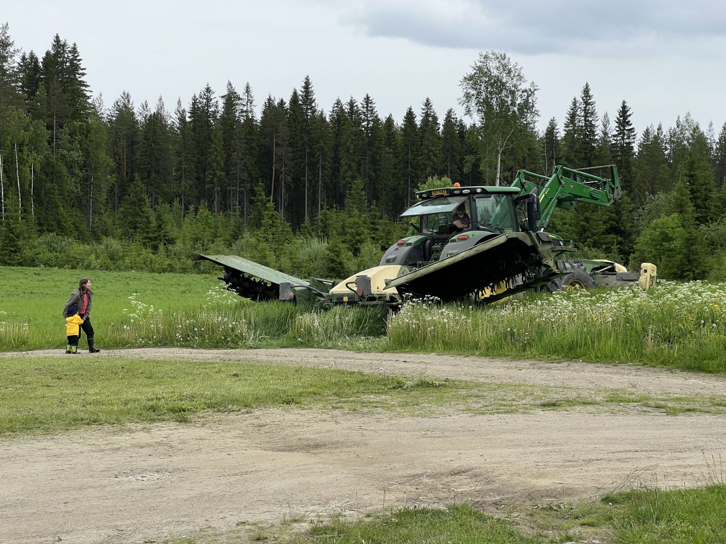 John Deeren iso vihreä traktori rehupellon laidalla aloittamassa heinän leikkaamista. Traktorin takaosa on näkyvissä, ja takana on kaksi isoa leikkurisiivekettä nostettuna maasta irti. Pellon edessä on hiekkatie, jossa nainen ja pieni lapsi katsovat traktoria. Traktorin kuljettaja on avannut kopin takaosan ikkunan ja on kääntynyt puhumaan naiselle ja lapselle.