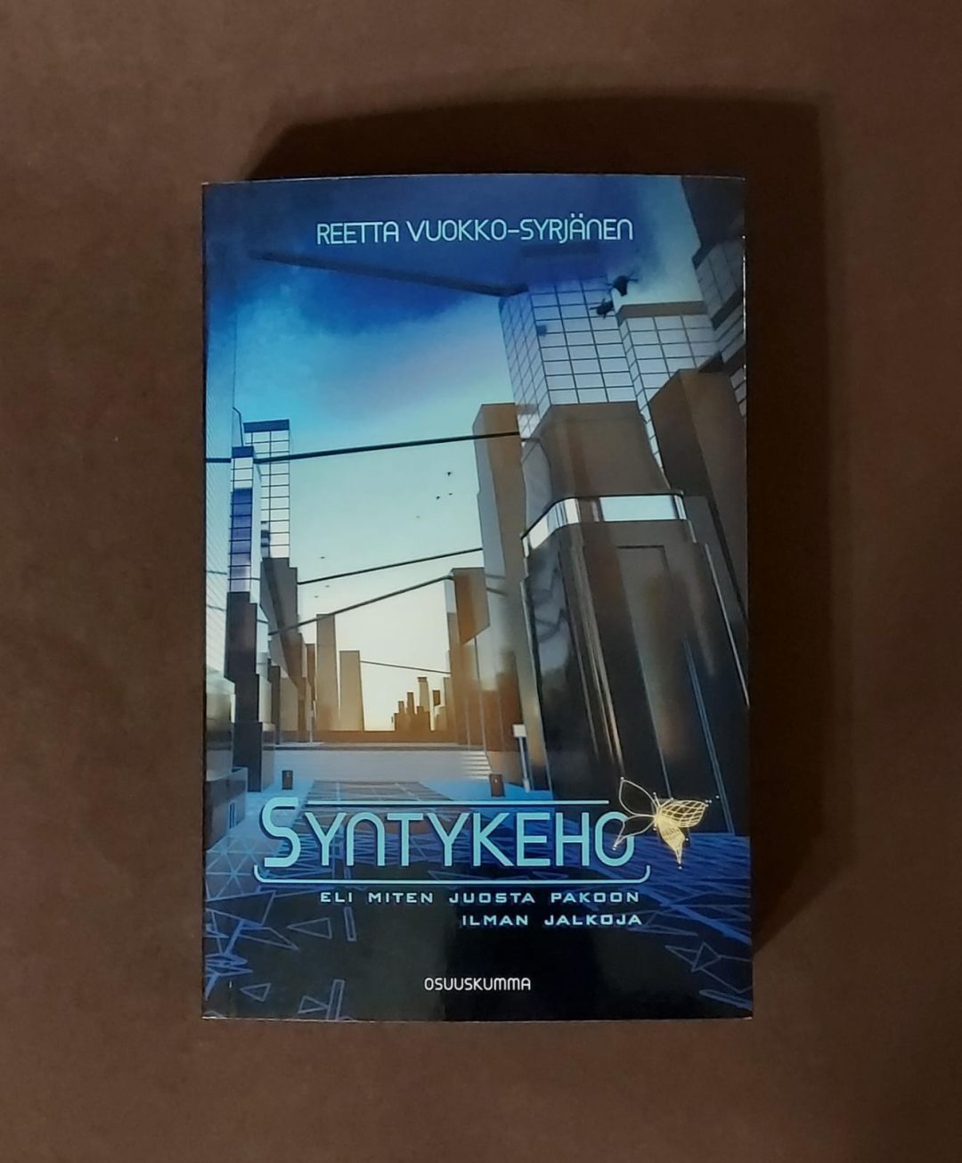 Kuvassa on kirja, jonka kannessa on futuristinen kuva sinisävyisestä suurkaupunkimaisemasta. Tie kulkee kahden korkean talorivistön välissä kohti portaita. Taivaanranta kuultaa horisontista keltaisena ja taivaalla leijailee mustia pikku droneja. Kirjan nimi Syntykeho lukee alaosassa ja sen vierellä on keltainen digitaalisesti luodun näköinen perhosen hahmo.