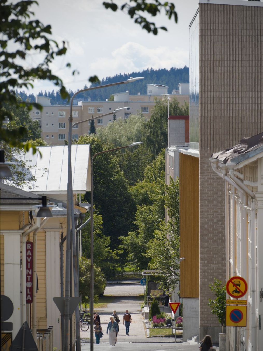 Valokuva kapeaa katua pitkin. Kadun varrella näkyy uusien ja vanhojen rakennusten sivuja ja joitakin käveleviä ihmisiä. Katu katoaa puiden sekaan keskellä kuvaa. Puiden takana näkyy kerrostaloja ja metsäistä mäkeä.