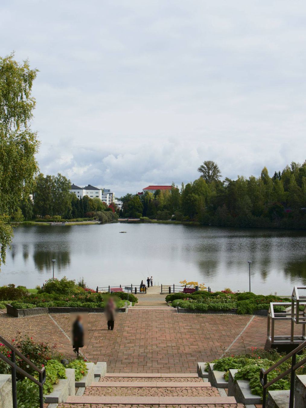 Finnish: Kuva portailta kohti järveä. Portaat laskeutuvat pienelle tasanteelle, jossa kävelee kaksi ihmistä. Ihmisiä sumennettu, etteivät ole tunnistettavissa. 

Tasanteen reunoilla kukkaistutuksia.

Portaat jatkuvat tasanteelta järven rantaan, jossa on kaksi poikaa onkimassa laiturilla.

Rantaa sivuaa polku ja katuvaloja. Vastarannalla näkyy järveä ympäröivä polki, metsää ja kerrostaloja. 

Keli on pilvinen ja ruskan merkkejä voi havaita tarkasti katsomalla. Puut ovat kuitenkin pääosin vihreitä.

English: Photo taken from stairs towards a lake. Stairs leads to a small square where two people are walking. They are blurred so they wouldn't be identified.

The square is surrounded by plantings.

Stairs leads from the square to the shore and there is two boys fishing on a small jetty.

On the opposite shore there is a path, forest and buildings.

Weather is cloudy and early signs of autumn can be seen. Trees are still mostly green.